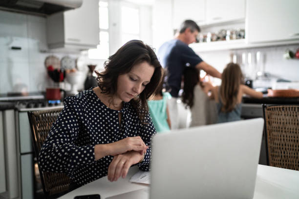 femme mûr travaillant vérifiant l’heure et utilisant l’ordinateur portatif avec la famille sur l’arrière-plan à la maison - vérifier lheure photos et images de collection