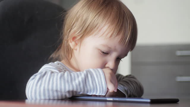 Portrait of toddler sitting at desk and tapping screen of digital tablet.