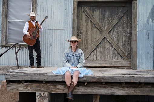 A cowboy step-father holding a guitar stands behind his cowgirl step-daughter, smiling, in blue dress, cowboy boots and hat, and denim jacket. They are in front of a rustic barn door in Texas. Copy space.