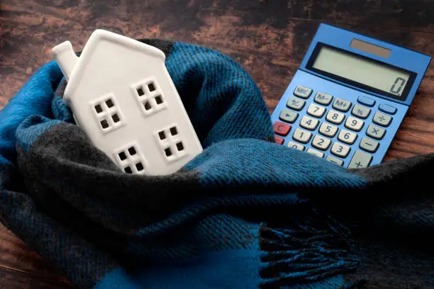 Photo of Warming efficiency of a residence and calculate heating costs concept with digital calculator, small house wrapped in warm wool neck scarf on wood background