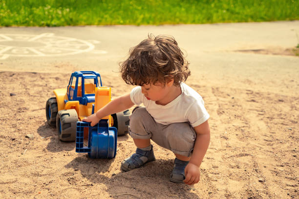 Little boy playing on the sand ground outdoors with a toy excavator Little long haired boy sitting on the sand ground outdoors and playing with a toy excavator sandbox photos stock pictures, royalty-free photos & images