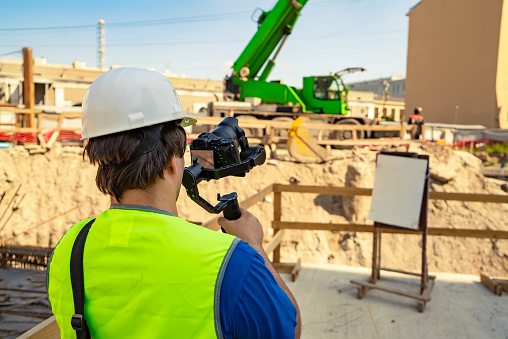 Videographer, dressed in safety gear, with a camera and stabilizer in his hands, filming a working process at a construction ground.