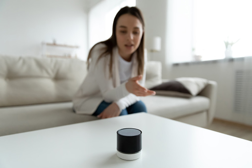Close up young woman talking to wireless smart speaker, sitting on couch at home, girl activating digital assistant, asking question to electronic device, searching in internet, listening to music