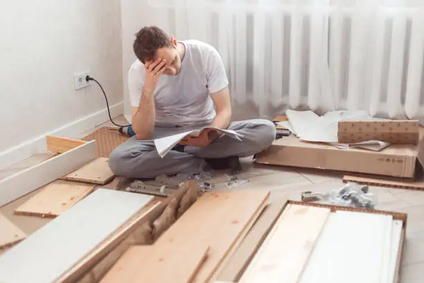 Bewildered man assembling wooden furniture. Difficulties and disadvantages of self-assembly of furniture at home without a master. Man reading instructions and misunderstand what to do the next.