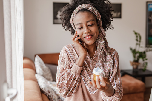 Una mujer afroamericana llama a los servicios de salud por teléfono y busca consejo médico mientras sostiene una botella de pastillas en la mano photo