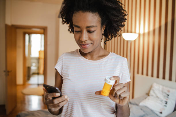 African American woman is looking for information on the internet about the medicine she is holding in her hand African American woman is sitting on the bed in the bedroom communicating with the doctor online while holding a bottle of medicine in her hand pill bottle photos stock pictures, royalty-free photos & images