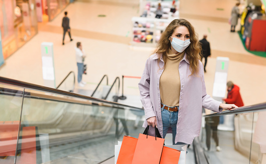 A young woman wearing a medical mask on an escalator in a shopping mall. A woman holding bags with clothes and shopping from different stores. Shopping in the new normality after Epidemic of Covid-19