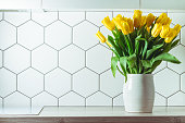 Interior shot. Yellow tulip bouquet in white vase on kitchen countertop on hexagonal white tile background. Copy space