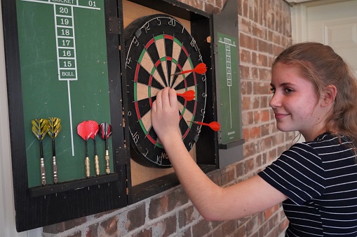 Pre-teen girl smiles as she counts her points and pulls darts from a dart board. Close-up, waist-up, profile.