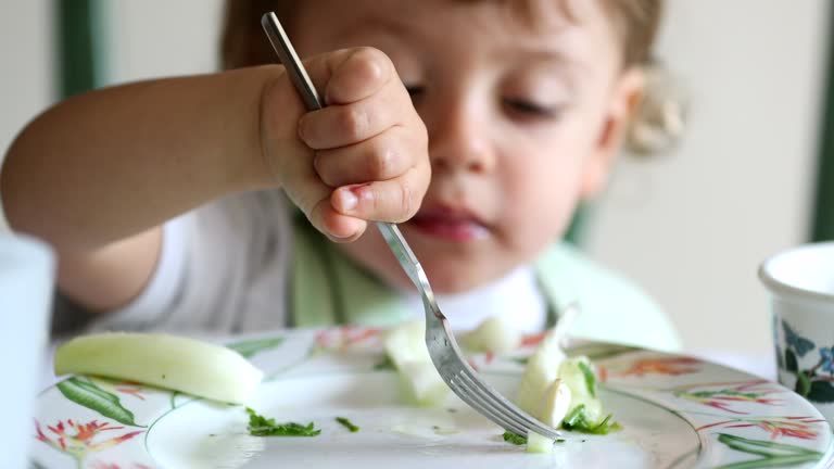 Child eating healthy vegetable with fork, close-up face baby toddler eats meal learning to use fork