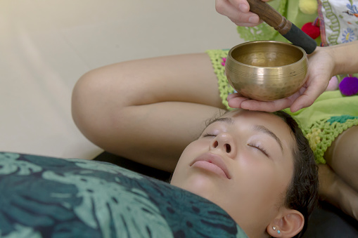 A single athletic middle age young active latin Spiritual Seeker woman practicing yoga and Meditating while playing on Tibetan singing bowl as a therapy to another young teenager woman inside a studio. The Professor is wearing a green Indian shirt as well the female young patient.

Sound therapy and meditation stock.