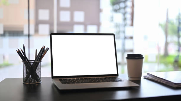 istock Mock up computer laptop with white screen, coffee cup, notebook and pencil holder on wooden table near window. 1311033277