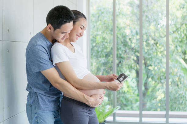 Mom and Dad are watching pictures of ultrasound in their hands. Young asian pregnant couple a child holding ultrasound. Mom and Dad are watching pictures of ultrasound in their hands. Young asian pregnant couple a child holding ultrasound. 3 months pregnant belly stock pictures, royalty-free photos & images