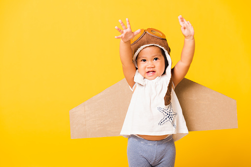 Happy Asian beautiful funny baby little girl smile wear pilot hat raise hand up play and goggles with toy cardboard airplane wings fly, studio shot isolated yellow background, Startup freedom concept