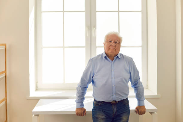 White-haired elderly man in shirt and eyeglasses looking at camera while leaning on table standing backlit near the window. Portrait of confident senior citizen, serious grandfather or house owner