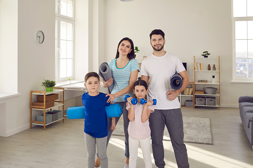 Active family sports workout in lockdown at home concept: Portrait of happy fit young couple with healthy little children standing together, holding exercise mats and dumbbells and looking at camera