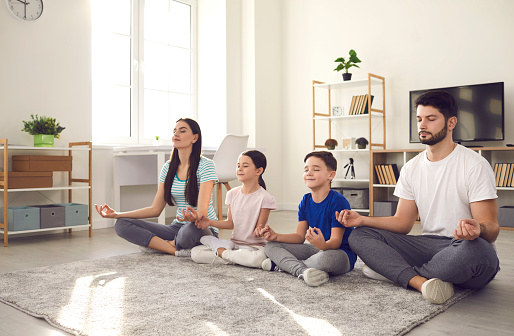 Healthy mind in healthy body. Millennial family with little children doing yoga at home. Happy young couple with calm kids sitting legs crossed on warm floor rug and practicing meditation together