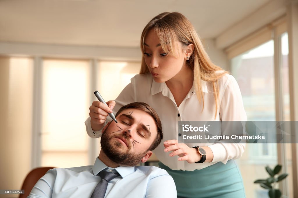 Young woman drawing on colleague's face while he sleeping in office. Funny joke Mischief Stock Photo