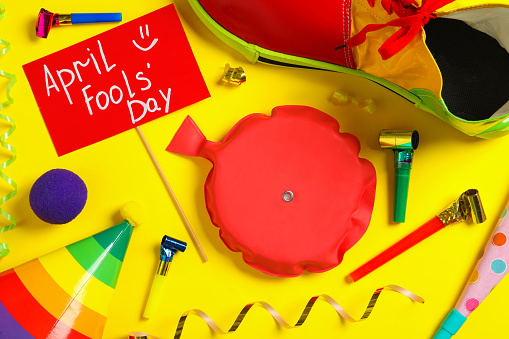 Sign with phrase April Fools' Day and different clown's accessories on yellow background, flat lay