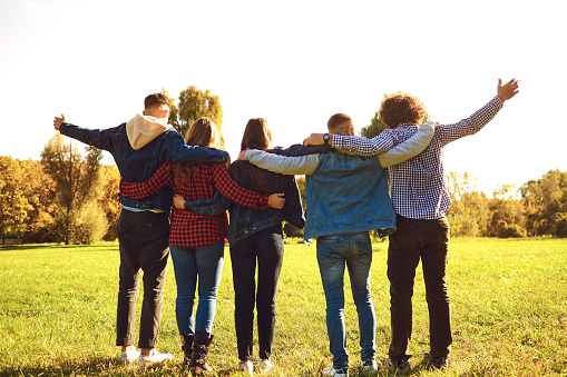 Back view of men and women embracing happily while standing on spacious green meadow in park having fun