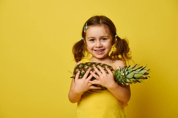 entzückende kleine mädchen, in gelb gekleidet, hält eine ananas und lächelnd niedlich, posiert vor der kamera auf einem gelben hintergrund mit kopierraum - facial expression isolated lifestyles exoticism stock-fotos und bilder