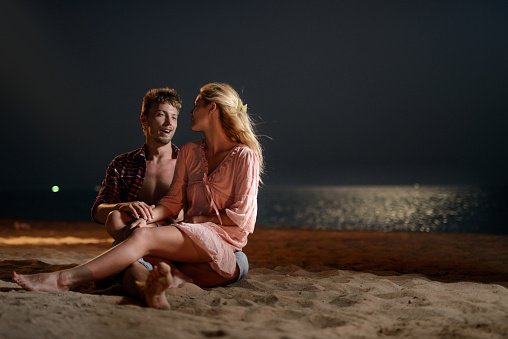 Young happy couple communicating while relaxing in sand on the beach at night. Copy space.