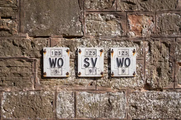 Photo of Row of ancient old English Victorian iron fire hydrant sign plaques on stone granite wall black and white. Symmetrical row of steel signs on rocks showing WO and SV