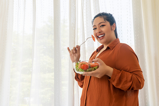 Concept food good healthy for weight loss. Smile obesity plus size women eat green organic vegetables while standing beside window in the room. Fat female holding glass bowl with salad vegetarian at home.