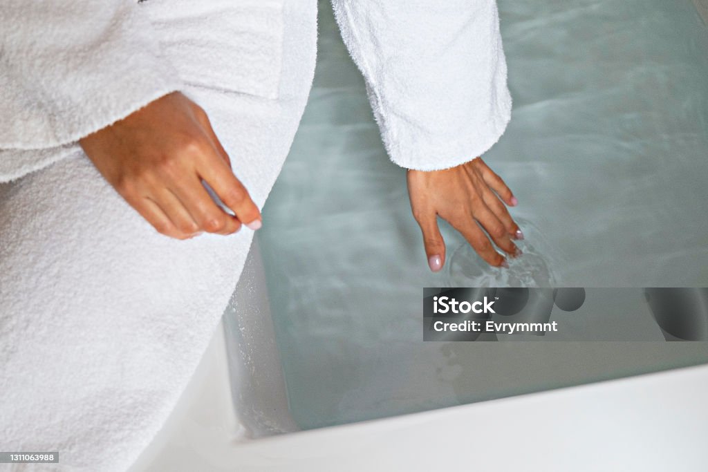 Young woman in a bathroom Cropped shot of young woman wearing the white bathrobe touching the water in a filled bathtub. Unrecognizable female testing the temperature of a tub with her fingers. Close up, copy space, background Bathtub Stock Photo