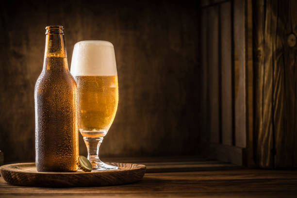 Beer bottle with a drinking glass full of beer on a rustic wooden table Front view of a beer bottle with a drinking glass full of beer on a rustic wooden table. At the lower side of the bottle is a golden cab. The objects are at the left side of the image leaving a useful copy space at the right side on the table. Predominant color is gold and brown. Low key DSLR photo taken with Canon EOS 6D Mark II and Canon EF 100 mm f/ 2.8 lager stock pictures, royalty-free photos & images