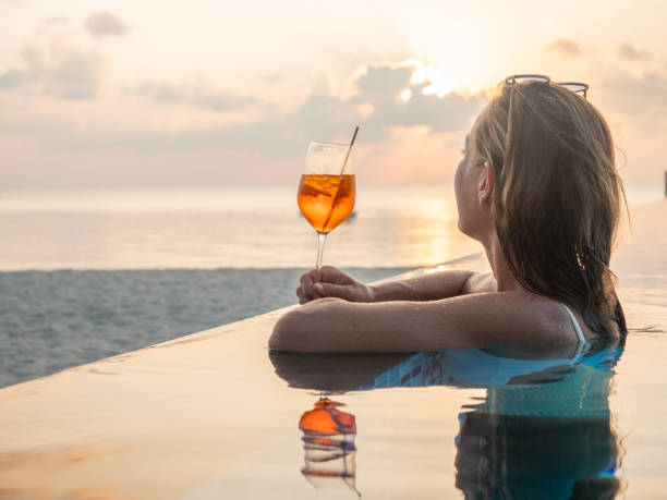 frau trinkt cocktail bei sonnenuntergang in einem infinity-pool - vacations infinity pool relaxation women stock-fotos und bilder