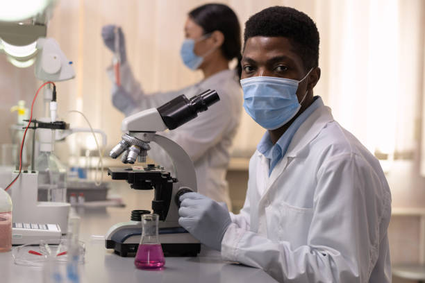 Chemist in mask working in the lab Portrait of African young chemist in mask looking at camera while working with microscope with his colleagues in the lab Pharmacy School stock pictures, royalty-free photos & images