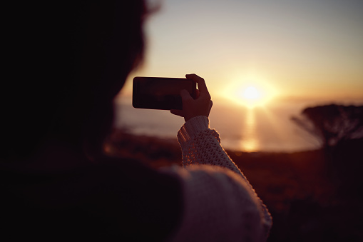 Shot of a woman taking pictures on her cellphone of the sunset