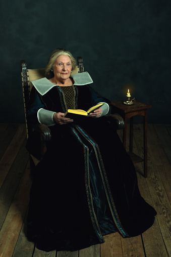 Mujer mayor vestida con atuendo renacentista con un libro sentado en una silla junto a una pequeña mesa rematada por una vela cerca de una pared oscura. photo