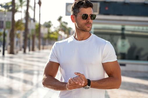 Portrait of handsome young man wearing sunglasses and white tshirt, posing on city street background. Casual style. Fashionable guy.
