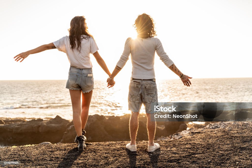 Beach couple watching sunset holding hands. Back view of man adn woman relaxing , enjoying summer. Beach couple watching sunset holding hands. Behind Stock Photo