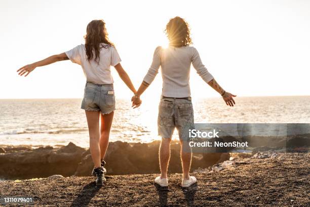 Beach Couple Watching Sunset Holding Hands Stock Photo - Download Image Now - Behind, Couple - Relationship, Young Couple