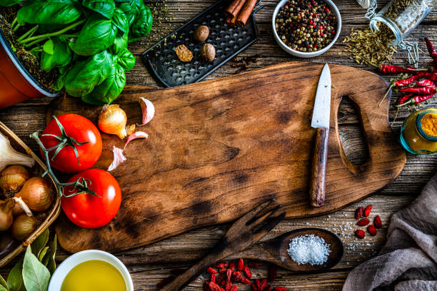 Kitchen and cooking backgrounds: overhead view of an empty cutting board with multi colored herb, spices and vegetables arranged all around the border making a frame shot on rustic wooden table. Useful copy space available for text and/or logo on the cutting board. High resolution 42Mp studio digital capture taken with SONY A7rII and Zeiss Batis 40mm F2.0 CF lens