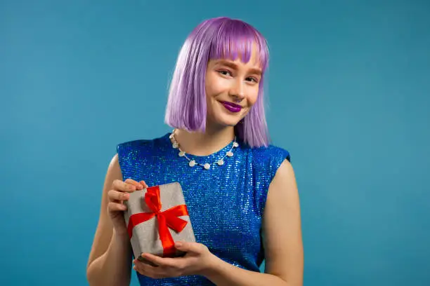 Excited trendy woman with violet hair received gift box with bow. She is happy and flattered by attention. Girl smiling with present on blue background. Studio portrait.