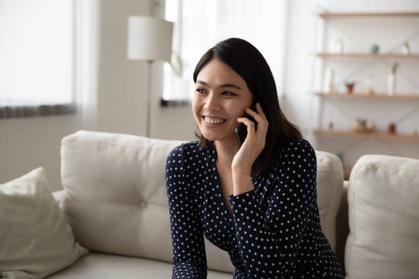 Smiling vietnamese female communicate with relatives abroad by roaming call Confident young asian lady spend time in pleasant phone conversation on sofa at living room. Smiling vietnamese female hold cell to ear communicate with relatives abroad by international roaming call answering stock pictures, royalty-free photos & images