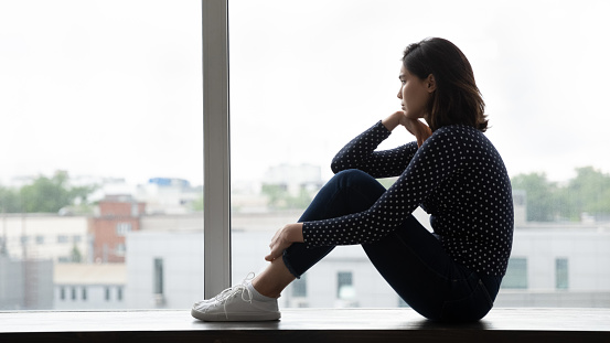 Side shot of pensive unhappy young lady of asian ethnicity sit on window sill hug knees look far make hard decision. Sad teen female think on unwanted pregnancy problem ponder on abortion. Copy space