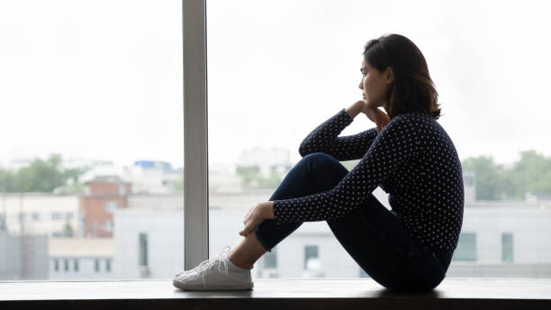 jovem asiática senta-se na janela abraça joelhos olhar para longe - human pregnancy pensive women thinking - fotografias e filmes do acervo