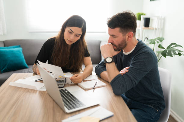 Stressed married couple checking their bills and debts Upset husband and wife doing their taxes with a laptop and bills in their living room. Latin young couple having financial and money problems bills and taxes stock pictures, royalty-free photos & images