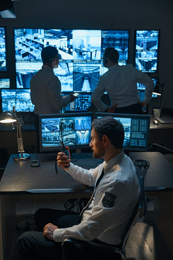 A man with a beard, a security guard or a police officer, possibly from the rescue service, is talking on a walkie-talkie against the background of digital monitors that display information from surveillance cameras. In the background, two of his colleagues stare intently at the security monitors.