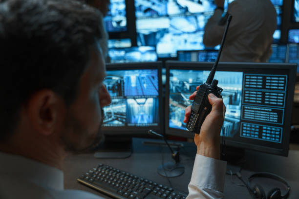 istock A security officer talks on a walkie talkie he is alone in the security room in complete darkness he is illuminated by monitors that display a picture from surveillance cameras 1311085375