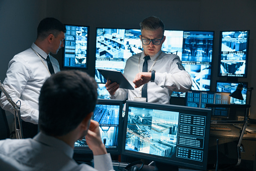 Security officers hold a meeting in the office of the central management and monitoring of the security object. The head of the security department looks at his watch, and checks the time with the rest of the security staff.