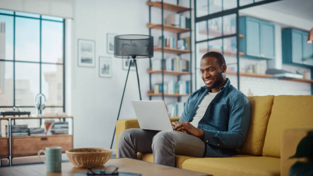 istock Handsome Black African American Man Working on Laptop Computer while Sitting on a Sofa in Cozy Living Room. Freelancer Working From Home. Browsing Internet, Using Social Networks, Having Fun in Flat. 1311087480