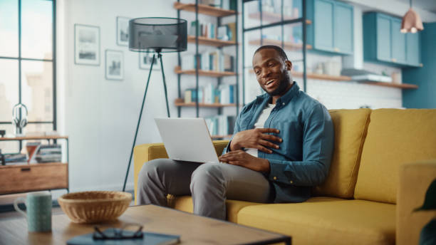 istock Handsome Black African American Man Having a Video Call on Laptop Computer while Sitting on a Sofa in Living Room. Freelancer Working From Home and Talking to Colleagues and Clients Over the Internet. 1311087482