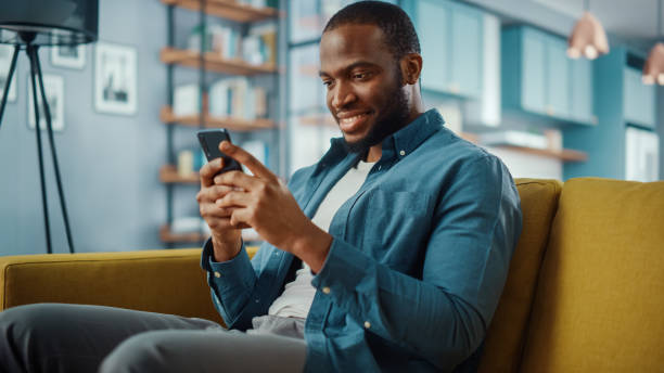 istock Handsome Black African American Man Using Smartphone while Sitting on a Sofa in Cozy Living Room. Freelancer Working From Home. Browsing Internet, Using Social Networks, Having Fun in Flat. 1311087486