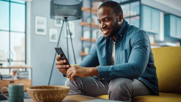 istock Handsome Black African American Man Using Smartphone while Sitting on a Sofa in Cozy Living Room. Freelancer Working From Home. Browsing Internet, Using Social Networks, Having Fun in Flat. 1311087487